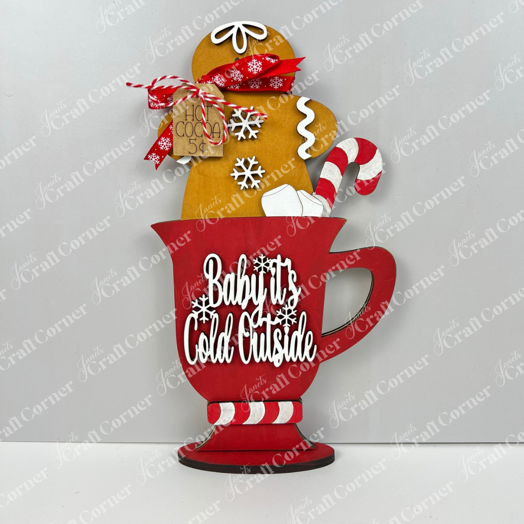 The Gingerbread Hot Cocoa Mug Shelf Sitter DIY kit from Janet's Craft Corner features a festive red mug with "Baby It's Cold Outside," gingerbread hot cocoa cookie, candy cane, snowflakes & tag—ideal for charming holiday shelf decor.