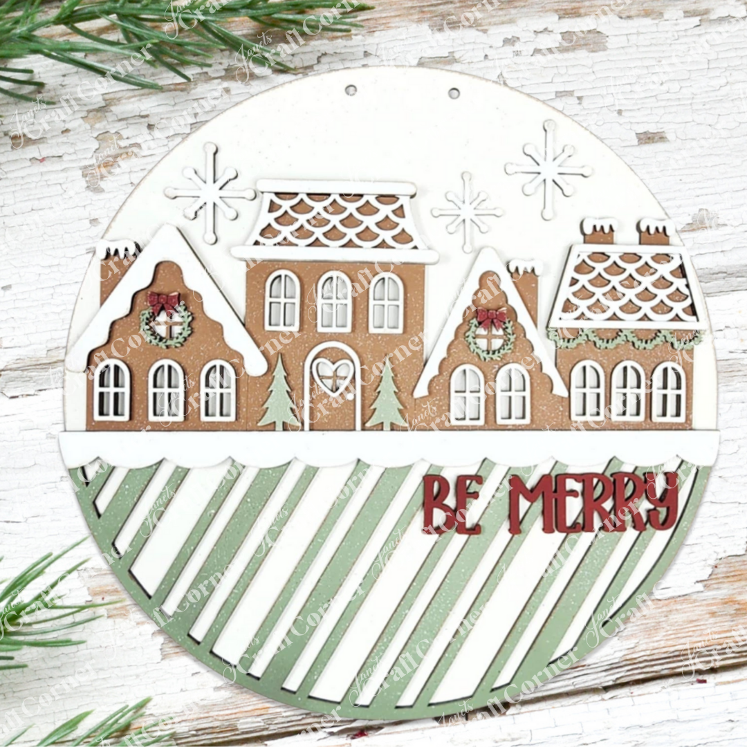 The Gingerbread Village Door Hanger by Janet's Craft Corner features gingerbread-style houses with wreaths, snowflakes, snowy rooftops, and "Be Merry" in red—ideal as festive décor or a DIY craft kit. Pine branches accent the corners for charm.