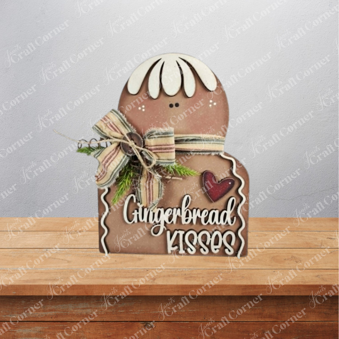 Add a festive touch to your home with the Gingerbread Kisses Shelf Sitter DIY home decor craft kit from Janet’s Craft Corner. This charming decoration features a gingerbread cookie design with white icing, a bow, greenery accents, and a heart, along with the phrase “Gingerbread Kisses.” Ideal as holiday shelf decor or as an adorable piece for your seasonal display, it comes in a set of 1 and is showcased on a wooden surface with subtle branding in the background.