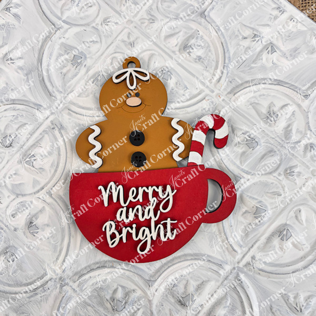 A smiling gingerbread figure sits in a red mug with a candy cane and "Merry and Bright" on the front—great holiday décor or inspiration for your next DIY holiday craft with Janet's Craft Corner's Gingerbread Ornaments kit (set of 8).