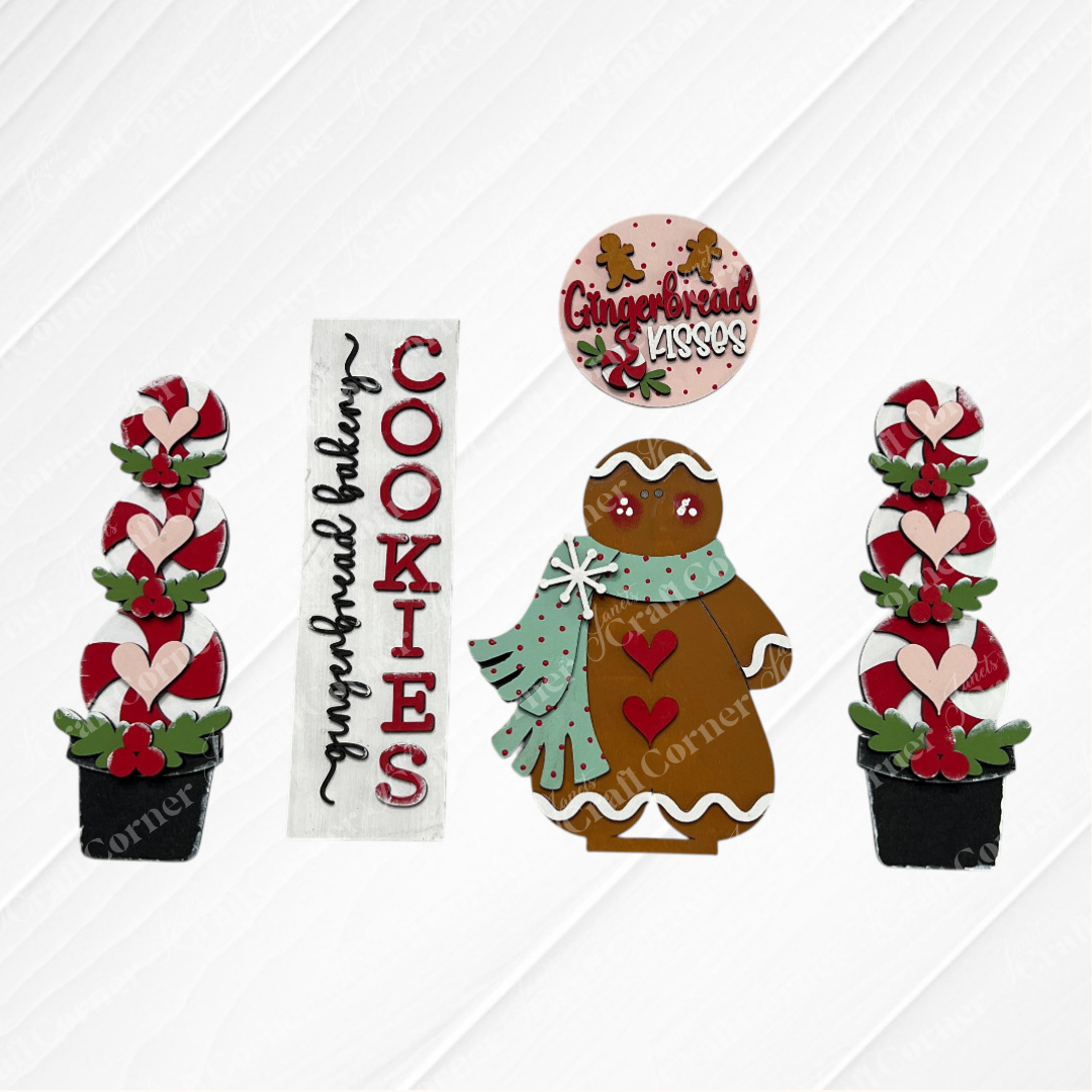 The Gingerbread Interchangeable Porch Set by Janet's Craft Corner includes six DIY seasonal decor pieces: a gingerbread person with scarf, “Cookies” sign, “Gingerbread Kisses” round sign, and two potted plants with red flowers and hearts.