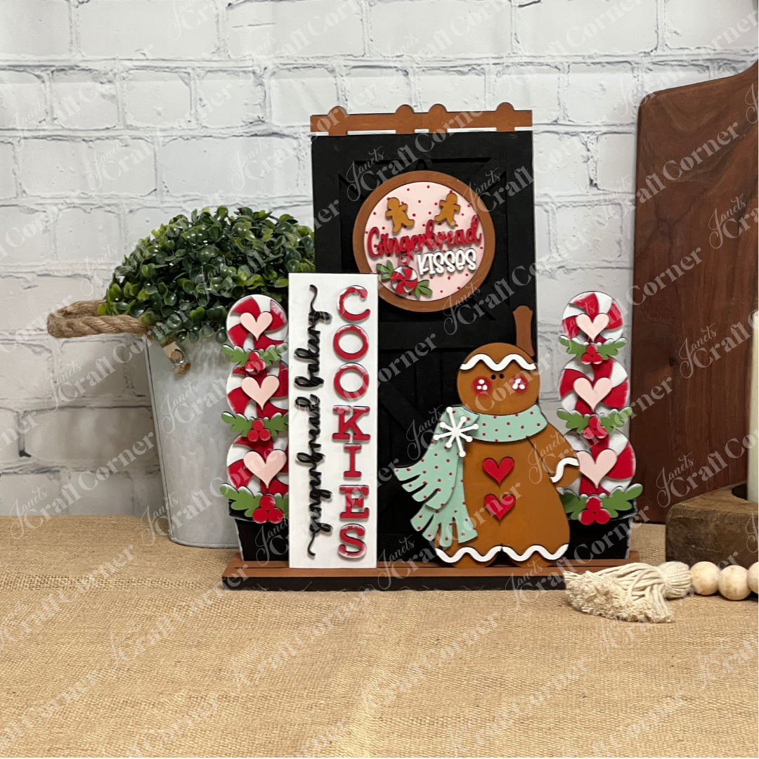 A festive display of Janet's Craft Corner Gingerbread Interchangeable Porch Set featuring a gingerbread cookie figure, "COOKIES" sign, heart accents, and a round "Gingerbread Kisses" sign on a burlap-covered table by a white brick wall.