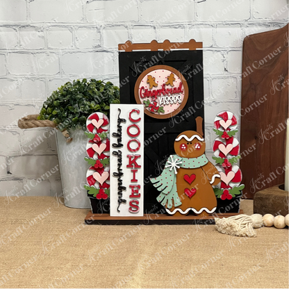 A festive display of Janet's Craft Corner Gingerbread Interchangeable Porch Set featuring a gingerbread cookie figure, "COOKIES" sign, heart accents, and a round "Gingerbread Kisses" sign on a burlap-covered table by a white brick wall.