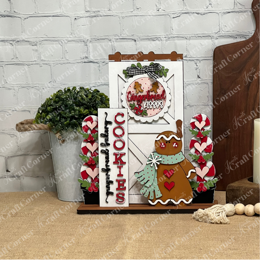 Janet's Craft Corner Farmhouse Porch Interchangeable Base DIY kit lets you create a festive gingerbread cookie display with a "Cookies" sign, candy canes, and a gingerbread figure—perfect for rustic farmhouse-style holiday decor.