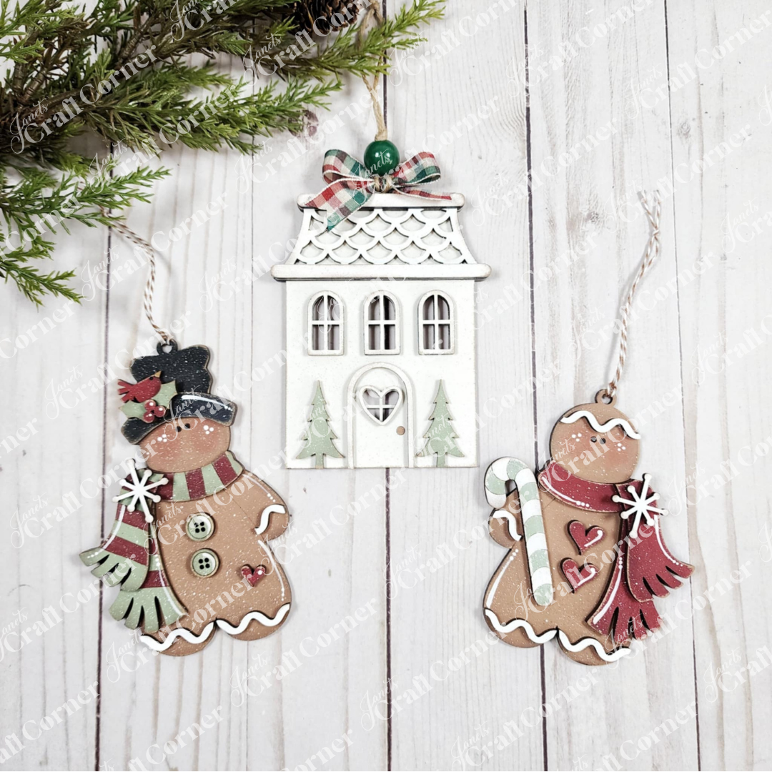 Three Christmas ornaments from Janet's Craft Corner's Gingerbread Ornaments DIY holiday craft kit (set of 8) rest on a white wooden surface, accented by pine branches for festive décor.