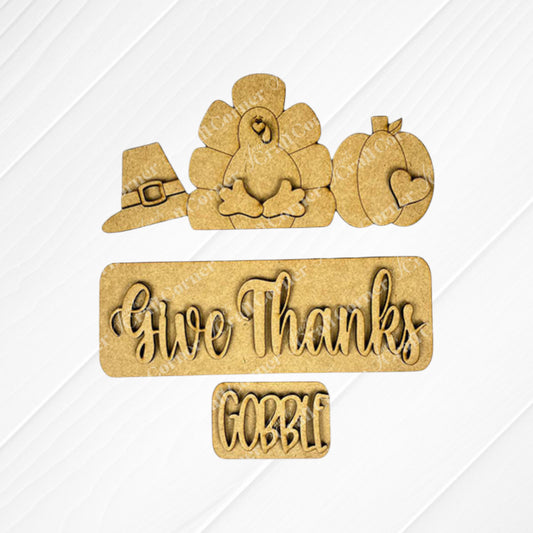 Enhance your home decor with Janet's Craft Corner's "Give Thanks Interchangeable Insert" DIY home decor kit. It features wooden cutouts showcasing a turkey, pilgrim hat, and pumpkin, along with the phrases "Give Thanks" and "Gobble," all set against a white background.
