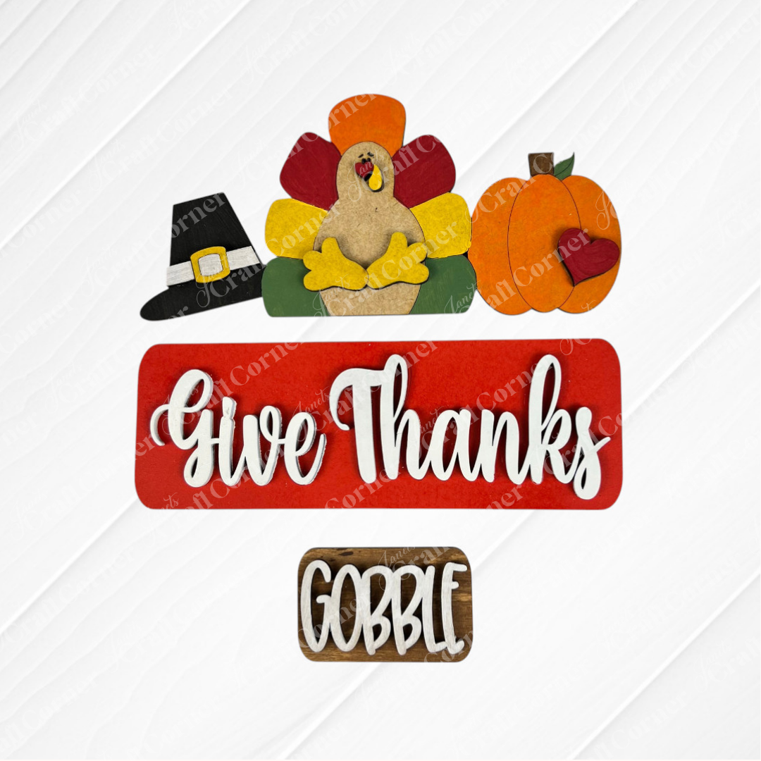 Janet's Craft Corner offers a festive DIY seasonal home decor kit featuring a cartoon turkey, pilgrim hat, and pumpkin with heart. A red sign says "Give Thanks," paired with a smaller wooden "Gobble" sign, adding charm to your space.