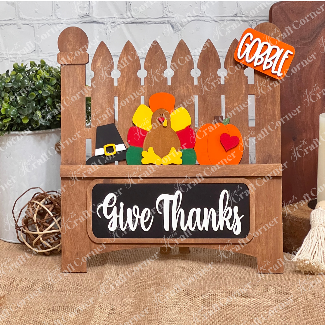 Janet's Craft Corner presents the Garden Fence with Insert, a DIY home decor kit featuring a white picket fence with a felt turkey, pilgrim hat, and pumpkin. "Gobble" adorns a top sign and "Give Thanks" graces the bottom plaque, ideal for crafting against any brick wall setting.