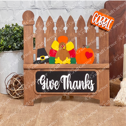 Janet's Craft Corner presents the Garden Fence with Insert, a DIY home decor kit featuring a white picket fence with a felt turkey, pilgrim hat, and pumpkin. "Gobble" adorns a top sign and "Give Thanks" graces the bottom plaque, ideal for crafting against any brick wall setting.