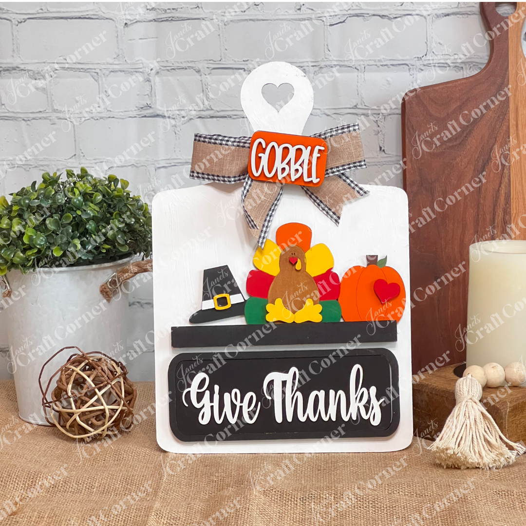 The "Breadboard Interchangeable Base with Insert" from Janet's Craft Corner features a turkey with colorful feathers, a pilgrim hat, and the words "Gobble" and "Give Thanks." This delightful DIY home decor piece rests against a white brick wall on burlap.