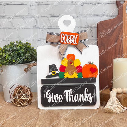 The "Breadboard Interchangeable Base with Insert" from Janet's Craft Corner features a turkey with colorful feathers, a pilgrim hat, and the words "Gobble" and "Give Thanks." This delightful DIY home decor piece rests against a white brick wall on burlap.