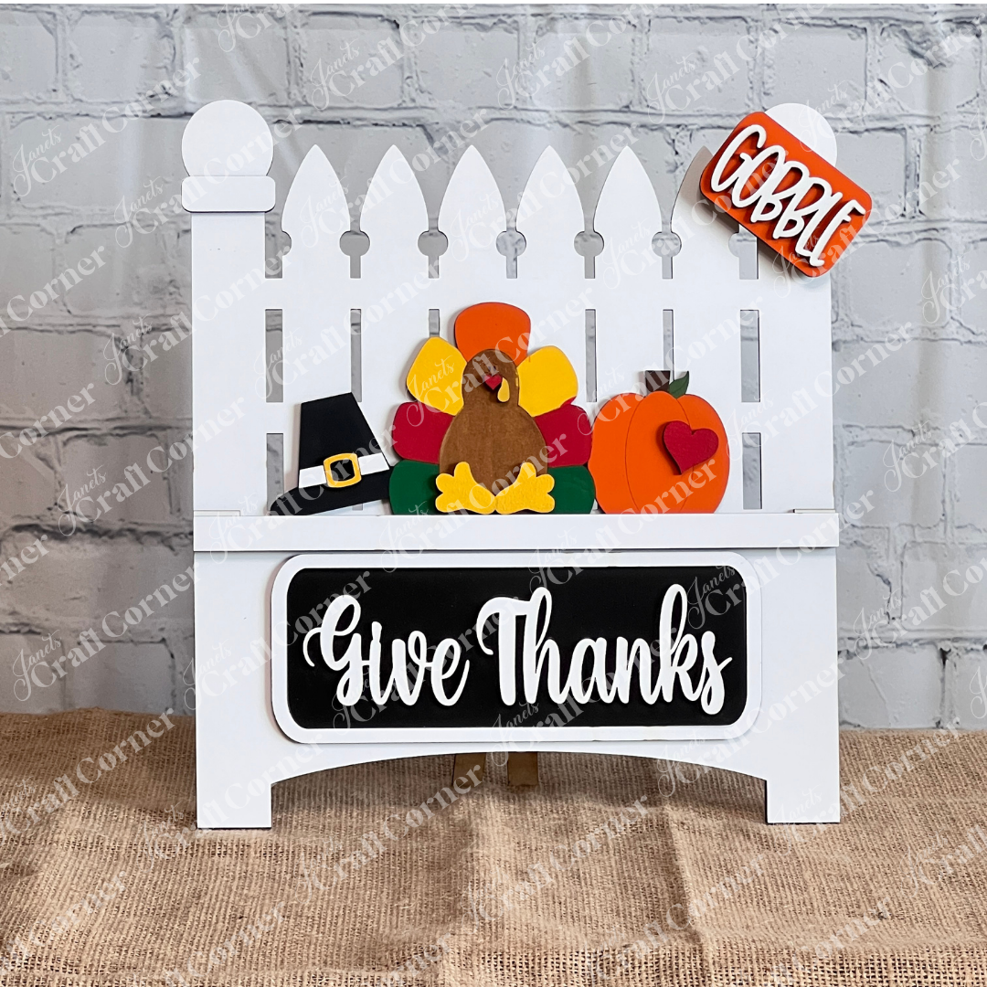 The "Give Thanks" interchangeable insert from Janet's Craft Corner is a DIY seasonal decor kit featuring a white fence with a turkey, pumpkin, and pilgrim hat. A sign reads "Gobble," and "Give Thanks" is elegantly displayed on a white garden fence against a brick wall.