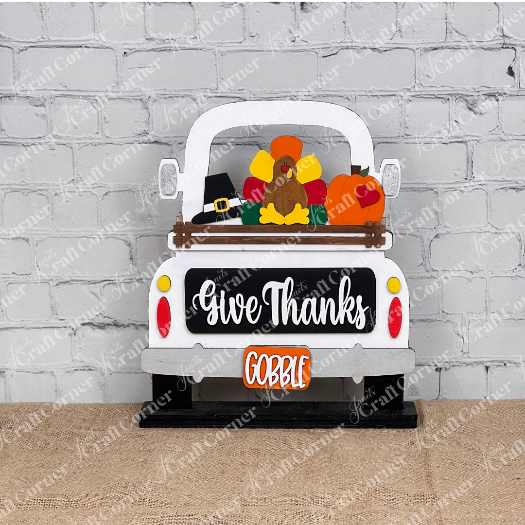 The "Give Thanks" Interchangeable Insert from Janet's Craft Corner showcases a wooden truck with turkey, pumpkin, and pilgrim hat elements. The words "Give Thanks" appear above "GOBBLE," ideal for DIY holiday decor against a brick wall.