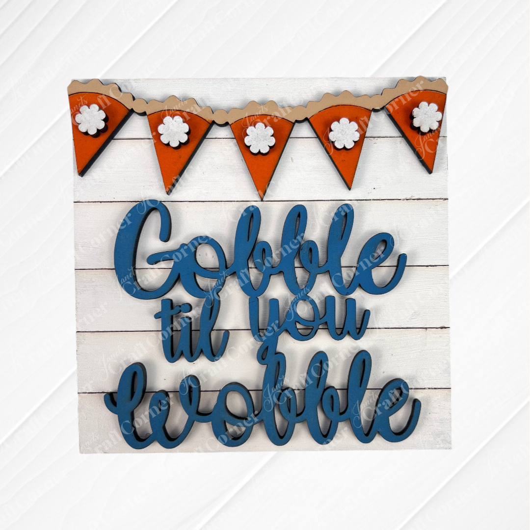 Janet's Craft Corner’s Thanksgiving Feast Squares feature blue cursive “Gobble til you wobble” on wood, with an orange floral pennant above—perfect tiered tray décor against a white wooden background.