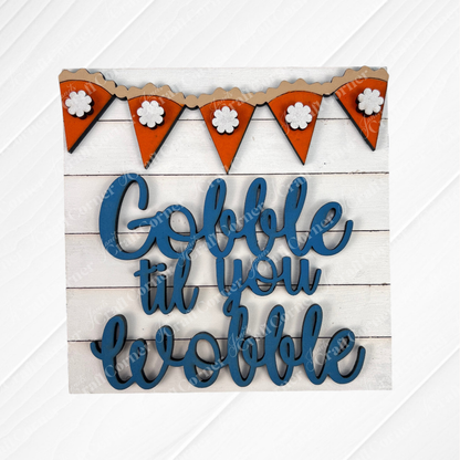 Janet's Craft Corner’s Thanksgiving Feast Squares feature blue cursive “Gobble til you wobble” on wood, with an orange floral pennant above—perfect tiered tray décor against a white wooden background.