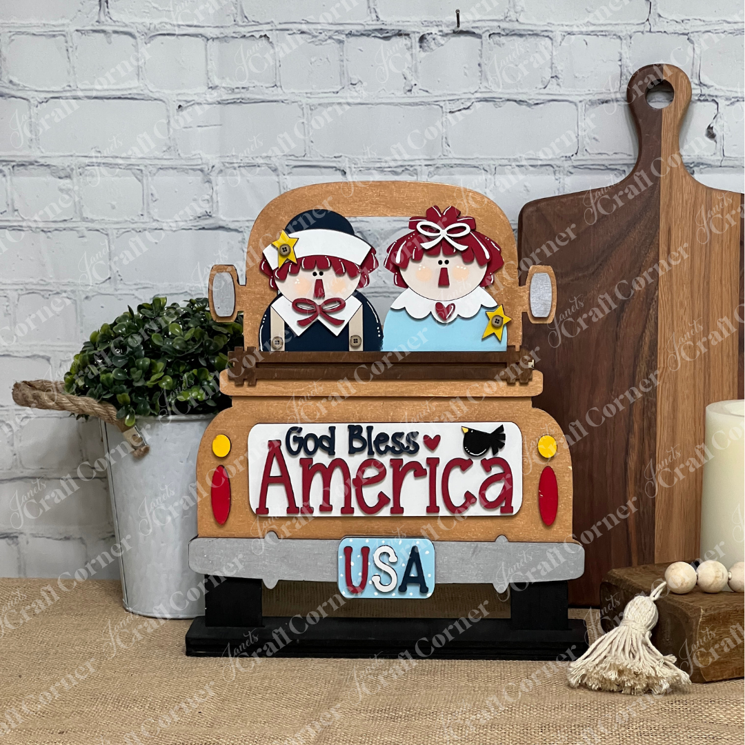 The God Bless America Interchangeable Insert from Janet's Craft Corner is a DIY seasonal decor kit featuring an unfinished wood truck with two dolls and patriotic sayings, perfect for display on a table with greenery and cutting boards.