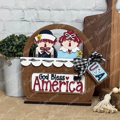 The God Bless America Interchangeable Insert DIY Craft Kit by Janet's Craft Corner includes 3 pieces with Americana dolls, a "God Bless America" message, plaid bow, "USA" tag, and patriotic accents—perfect for seasonal home decor.