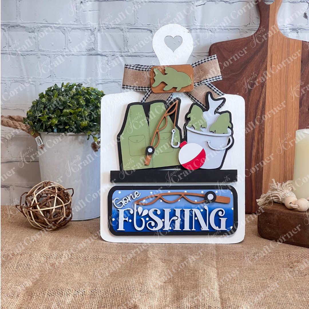 The Gone Fishing Interchangeable Insert from Janet's Craft Corner is a DIY seasonal home decor kit with 3 wood cut-out pieces: a green vest, fishing rod, and bobber over a "Gone Fishing" sign—ideal for craft enthusiasts.