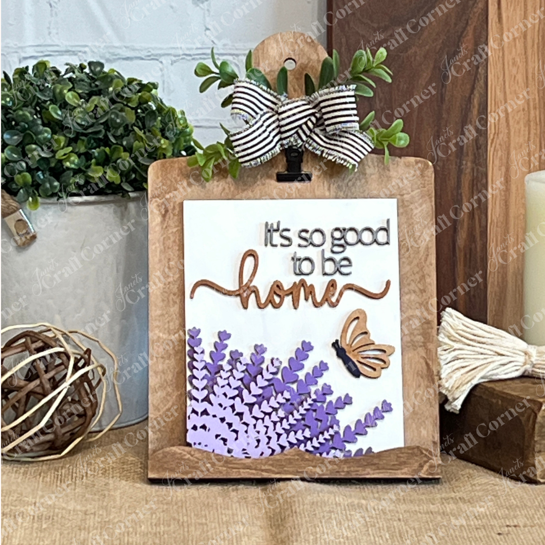 Janet's Craft Corner’s Lavender Switchable Farmhouse Signs DIY Craft Kit features a wooden stand displaying a lavender sign with flowers, butterfly, and “It’s so good to be home,” plus striped bow and greenery—perfect for rustic tiered tray décor.