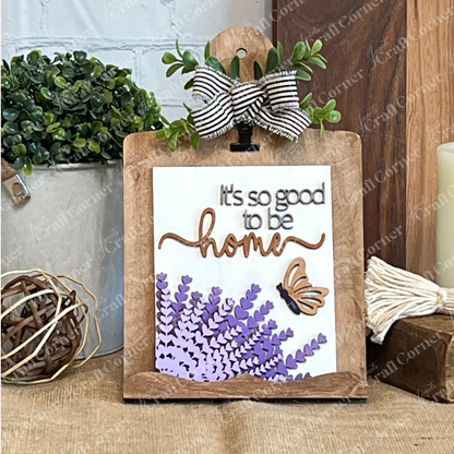 Janet's Craft Corner’s Lavender Switchable Farmhouse Signs DIY Craft Kit features a wooden stand displaying a lavender sign with flowers, butterfly, and “It’s so good to be home,” plus striped bow and greenery—perfect for rustic tiered tray décor.