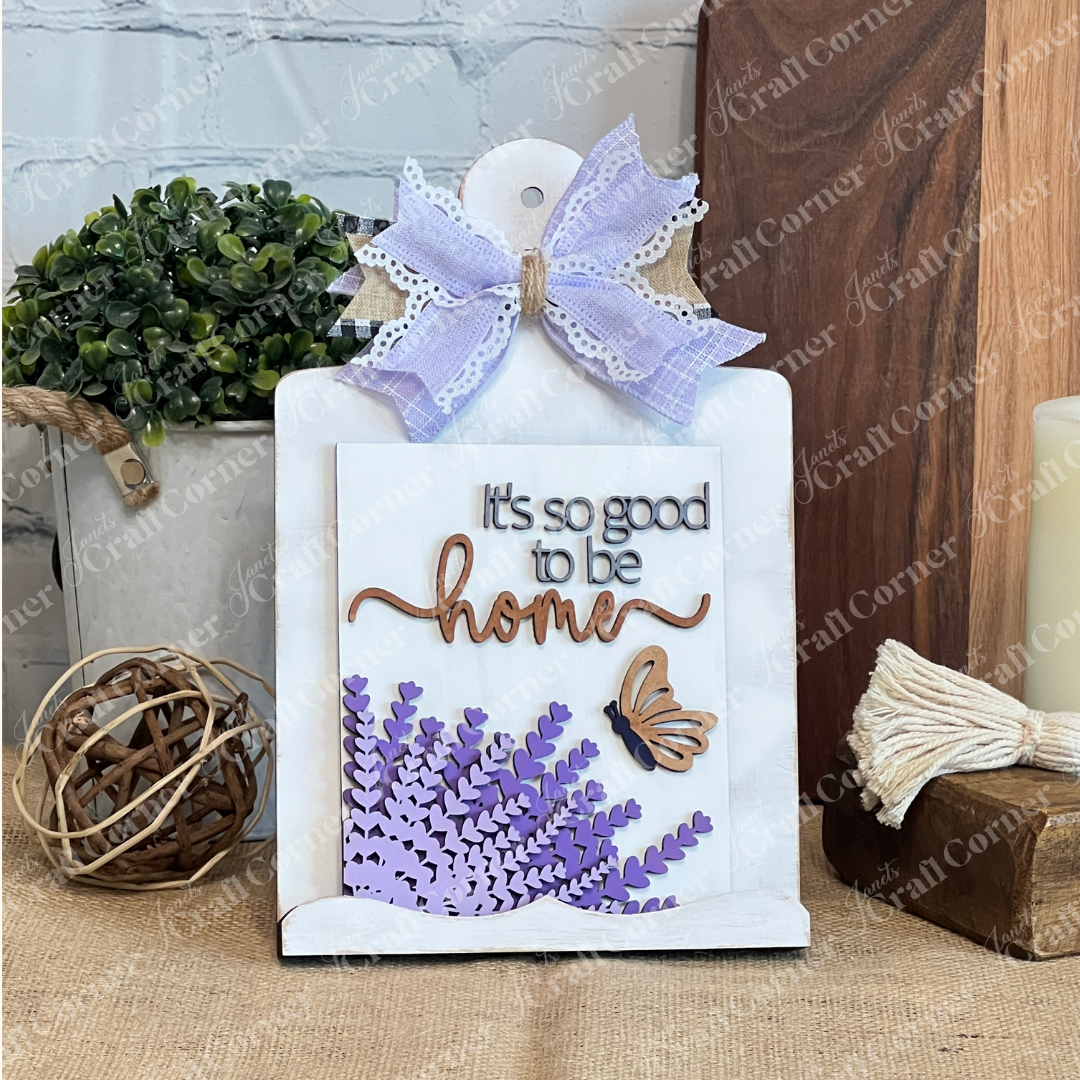Janet's Craft Corner’s Lavender Switchable Farmhouse Signs DIY Craft Kit features a butterfly and bouquet, reading "It's so good to be home." Ideal for tiered tray décor, it pairs beautifully with plants, baskets, or candles in any cozy space.