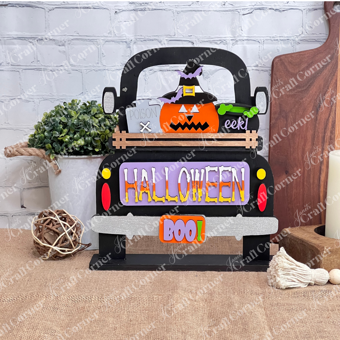 This delightful DIY home decor kit features Janet's Craft Corner's Halloween Pumpkin Interchangeable Insert, showcasing a truck with a cheerful pumpkin wearing a witch hat. The vibrant "HALLOWEEN" and "BOO!" text enhance its festive appeal. Accompanied by a plant, candle, and cutting boards set against a brick wall backdrop, it's ideal for those who enjoy interchangeable craft inserts.