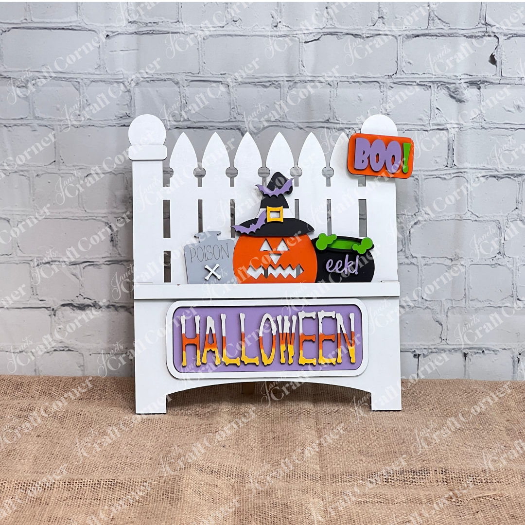This delightful Halloween decor from Janet's Craft Corner features a wooden fence backdrop with interchangeable inserts: a pumpkin, witch's hat, "Poison" bottle, and bubbling cauldron. A cheerful "BOO!" sign hangs above, while vibrant letters spell "HALLOWEEN" at the base.