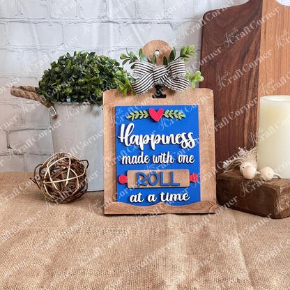 Janet's Craft Corner Kitchen Switchable Farmhouse Sign DIY kit features a wooden sign with "Happiness made with one roll at a time," bow, greenery, and paper roll art—perfect rustic decor for your kitchen. Set of 1.