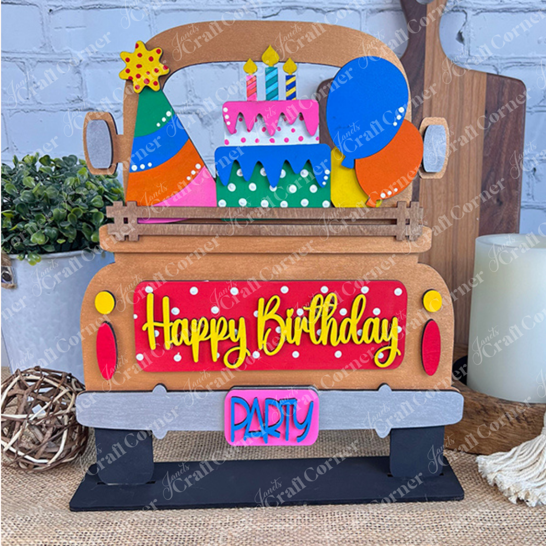 The DIY home decor kit from Janet's Craft Corner includes a wooden truck decoration featuring a "Birthday Interchangeable Insert." It is adorned with colorful balloons, cake, and party hats and is designed to stand among a small plant, candle, and wall decor to enhance any celebration.