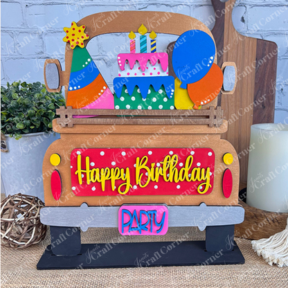 Introducing Janet's Craft Corner's DIY Home Decor Kit: The Antique Truck Base with Interchangeable Insert. This lively wooden truck display proudly showcases "Happy Birthday" on the back, complete with a party hat, balloons, and a layered cake design, perfectly suited for your festive occasions. It features an antique-style truck base that allows for customization with interchangeable inserts and is beautifully set against a rustic background alongside a small potted plant and charming decorative items.