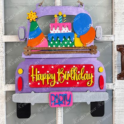 The "Birthday Interchangeable Insert" from Janet's Craft Corner features a vibrant wooden design of a truck with "Happy Birthday" on its tailgate, carrying a cake, balloons, and party hats. This DIY home decor kit is designed to hang on a white window frame against a brick wall for a festive touch.