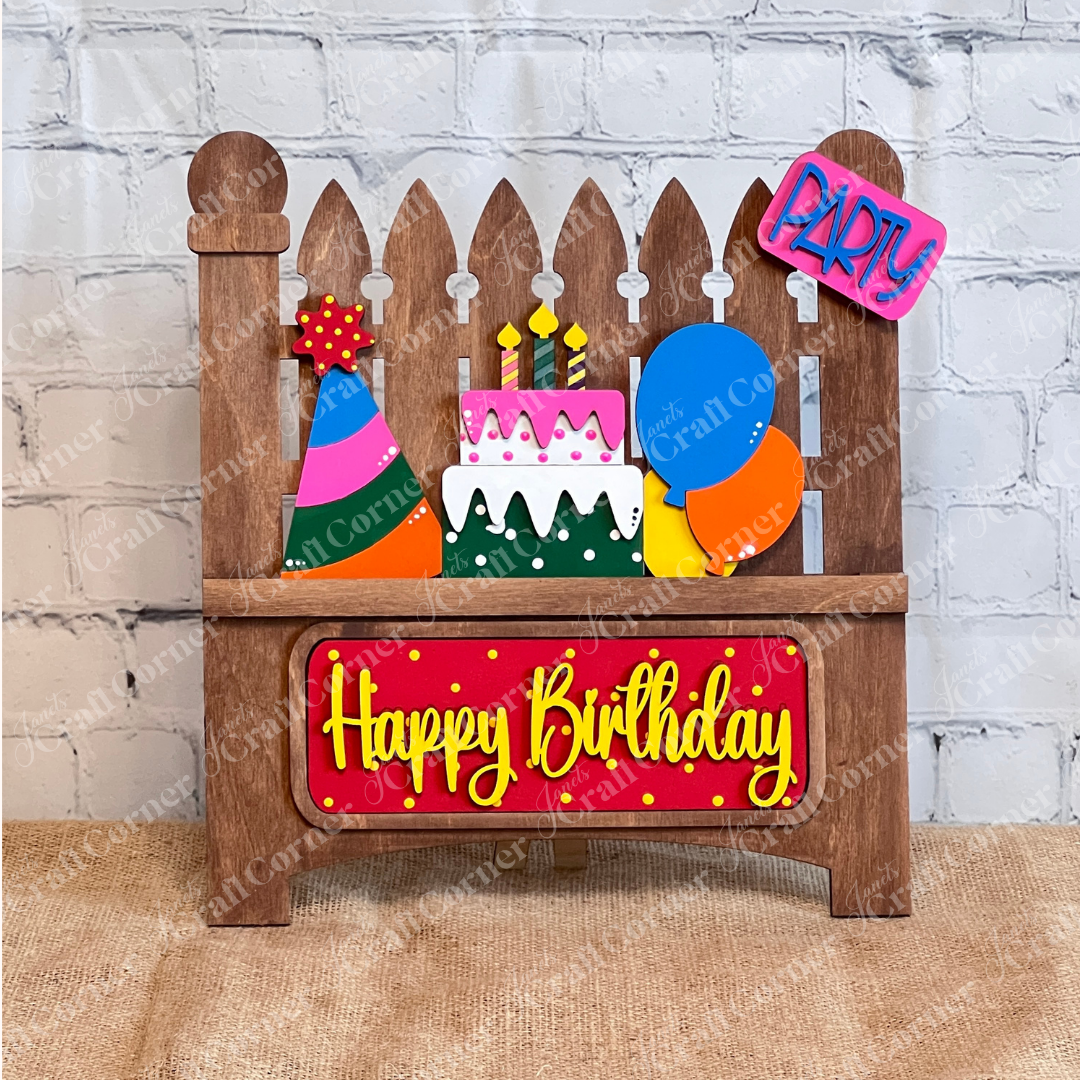 The Birthday Interchangeable Insert by Janet's Craft Corner features a wooden cake, candles, party hat, and balloons with "Happy Birthday" in yellow on a red background. Perfect for any birthday celebration, this DIY home decor craft kit includes a 3-piece set on a stained garden fence shelf sitter ideal for burlap surfaces against white brick walls.