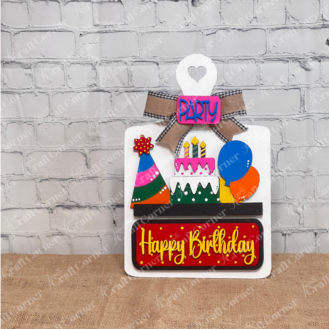 The Breadboard Interchangeable Base with Insert by Janet's Craft Corner is a DIY home decor kit featuring a wooden birthday theme with colorful elements like a cake and balloons, complete with "Happy Birthday" and "Party" signs, plus interchangeable accessories for personalized décor.