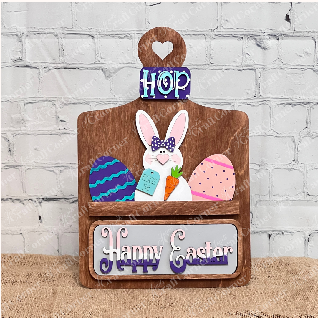 The Breadboard Interchangeable Base with Insert by Janet's Craft Corner is a DIY home decor kit featuring a wooden Easter sign: "HOP" at the top, a bunny with a carrot, surrounded by colorful eggs. Personalize it on a burlap-textured brick wall backdrop with "Happy Easter" charmingly at the bottom.