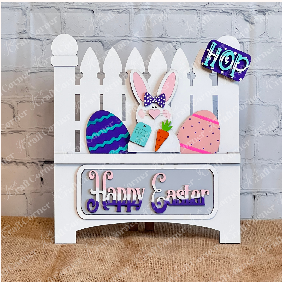 Add charm to your craft kit with Janet's Craft Corner DIY decor: a white picket fence featuring a rabbit with carrot, "EGGS 5¢" sign, colorful eggs, and "Hop" tag. Below, "Happy Easter" in purple and pink against a burlap and brick wall backdrop. Set includes 1 fence base and 1 insert.