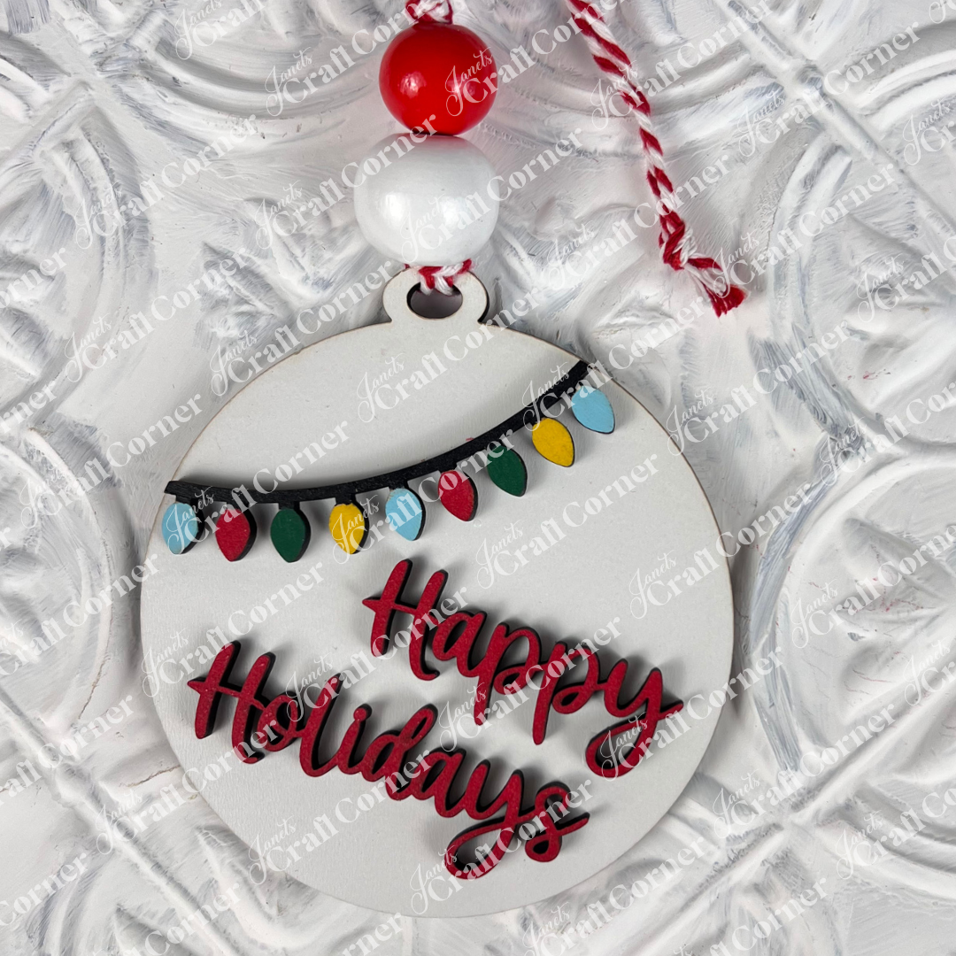 Introducing the Round Christmas Ornaments DIY Decoration Kit from Janet's Craft Corner—featuring a festive ornament with "Happy Holidays" elegantly scripted in red. Accentuated by a string of colorful lights, it hangs beautifully from a coordinating red and white cord with matching beads, enhancing any Christmas display against its textured white background.