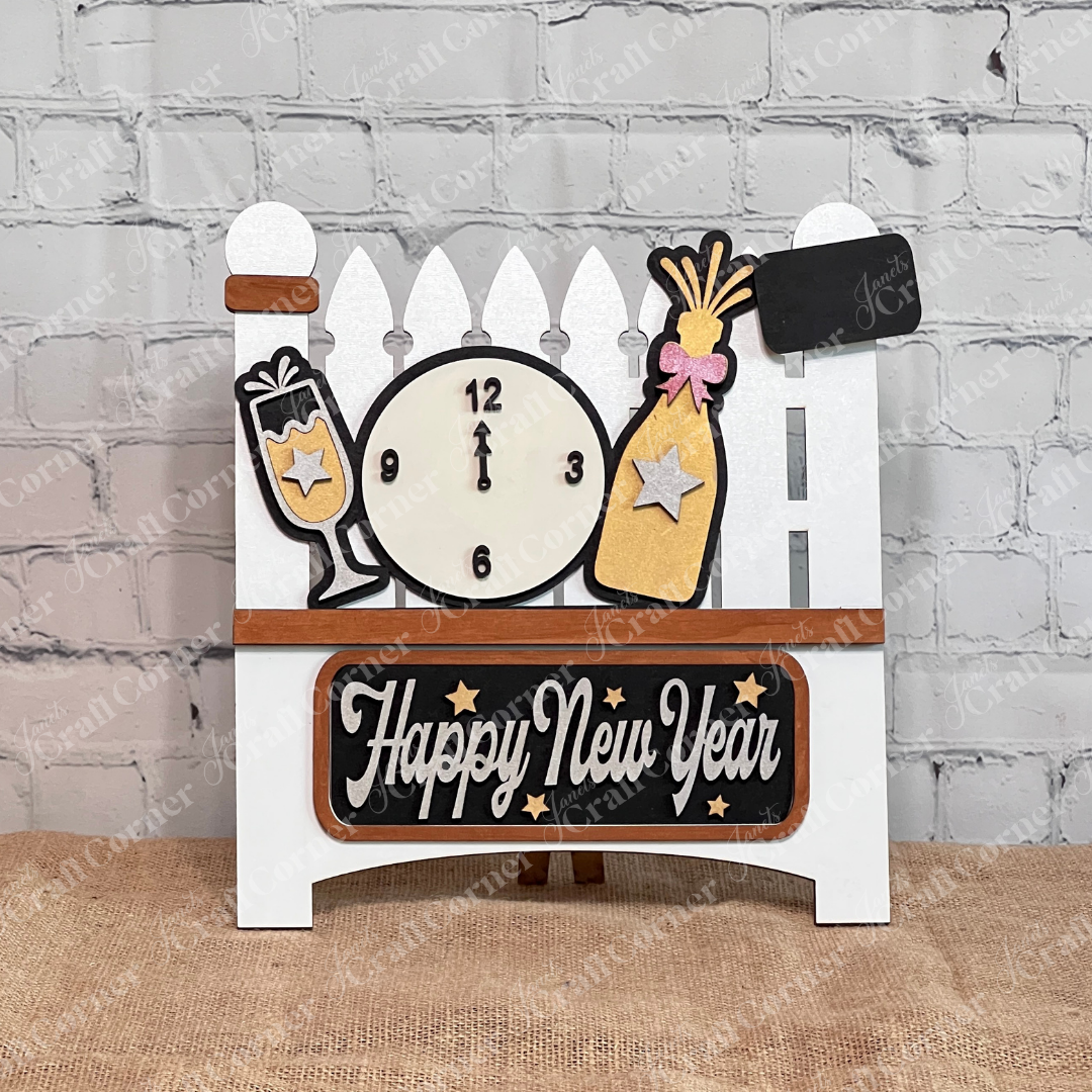 Janet's Craft Corner offers the New Year Interchangeable Insert DIY Kit on a white garden fence, featuring a clock at midnight, champagne elements in black and gold, "Happy New Year" script, and background decor with a plant, candle, and wooden board. Perfect for seasonal home decoration.