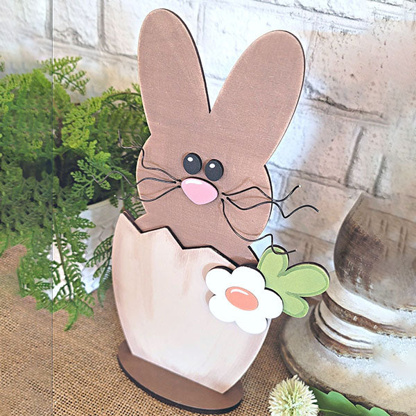 The Hatched Bunny Shelf Sitter from Janet's Craft Corner is a delightful wooden decoration. It showcases a brown Easter bunny with oversized ears and a pink nose peeking out from a cracked egg, embellished with a flower and green leaves. Ideal for home decor, it enhances rustic charm when placed against brick or greenery backgrounds.