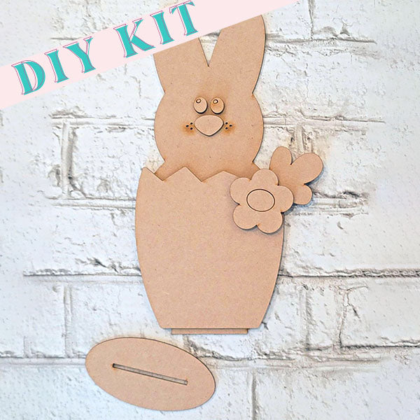 This DIY kit from Janet's Craft Corner is perfect for home decor and includes a Hatched Bunny Shelf Sitter, crafted from brown cardboard to resemble a bunny emerging from a cracked egg. A cut-out flower adorns the scene, set against a textured white brick wall backdrop.