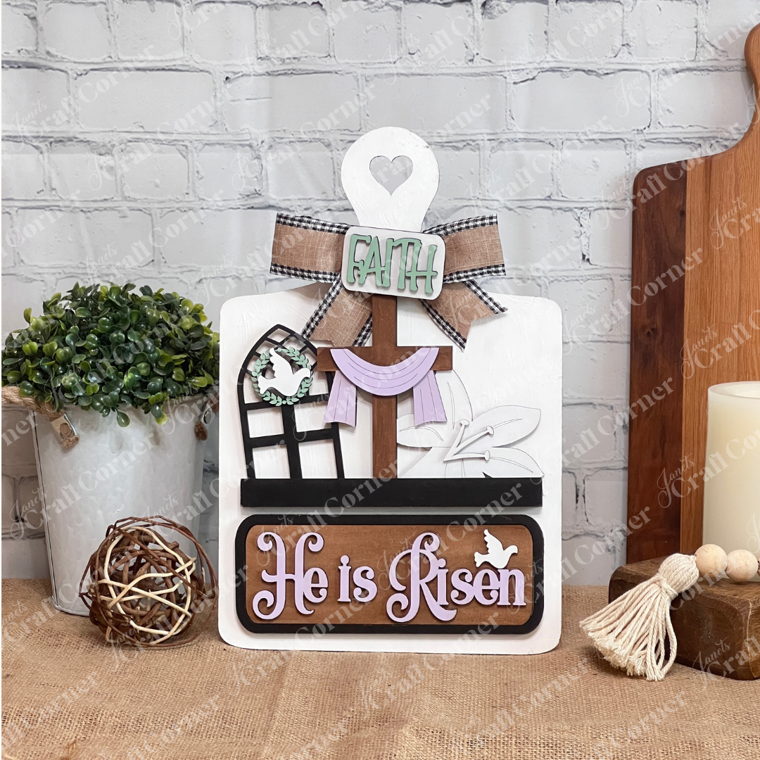 Janet's Craft Corner offers a DIY home decor kit featuring a breadboard base and insert with a cross draped in cloth, window silhouette with dove, and "He is Risen" in lilac text topped with "Faith," perfect for adding your personal touch against a white brick wall.