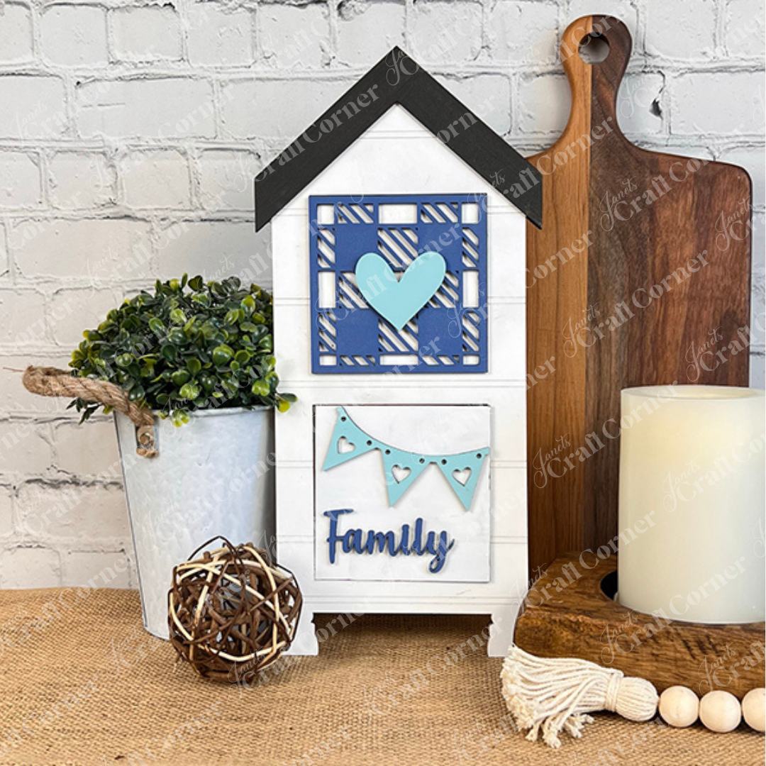 The Family Home Squares sign by Janet's Craft Corner, featuring a blue heart and "Family" text, is displayed on a table with tiered tray decor, a potted plant, wooden cutting boards, a candle, and twine ball against a white brick wall.