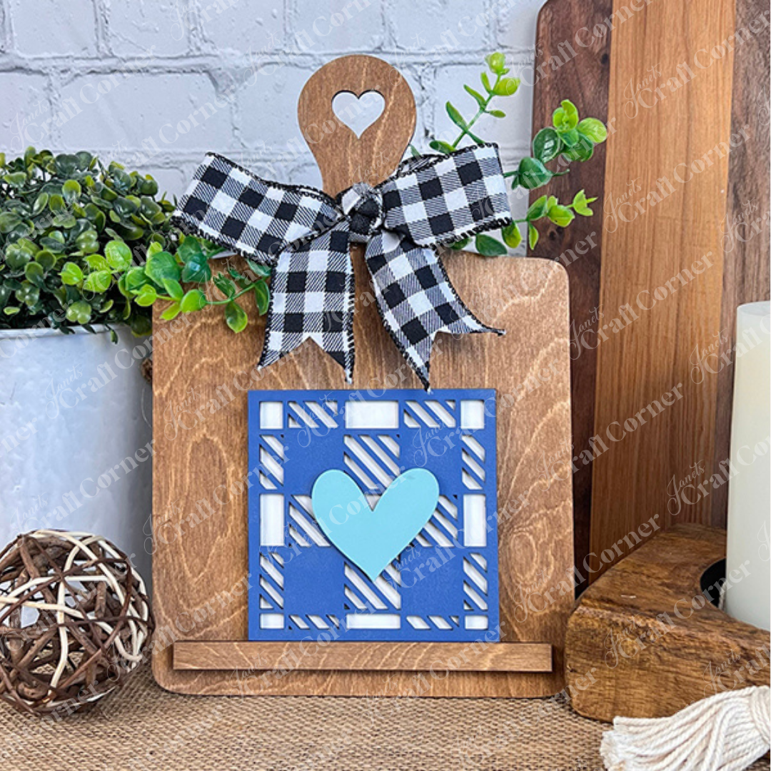 A wooden cutting board with a heart cutout and plaid bow displays the Family Home Squares by Janet's Craft Corner. Greenery and tiered tray decor surround it on the table, creating a cozy display.