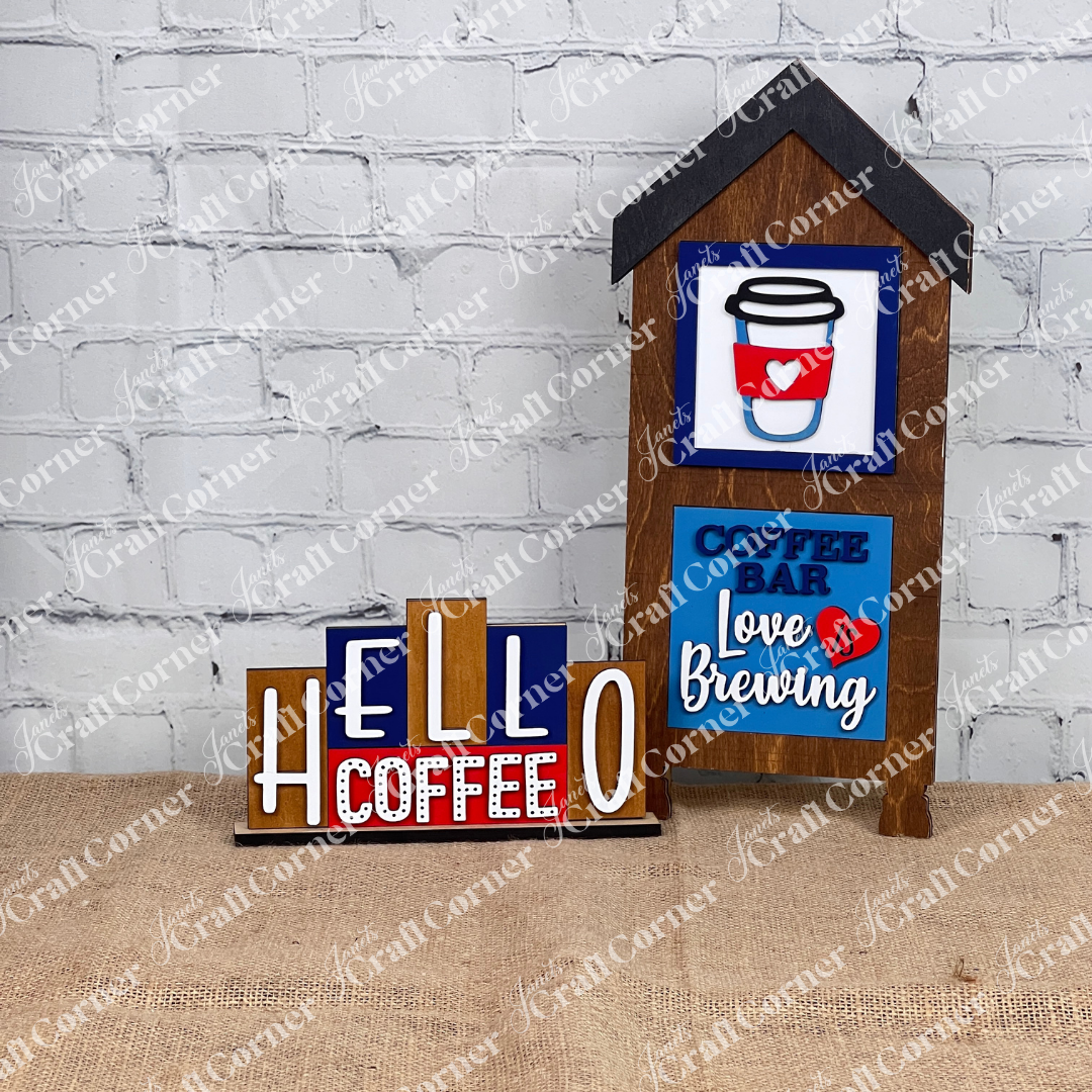 Set against a burlap backdrop and brick wall, the Decorative Coffee Signs by Janet's Craft Corner feature charming designs. The Hello Coffee Mini Word Block displays "HELLO COFFEE" in bold letters, while another sign, shaped like a quaint house, adds charm with phrases "COFFEE BAR" and "Love is Brewing," making it an ideal addition for any DIY kit enthusiast's home decor.