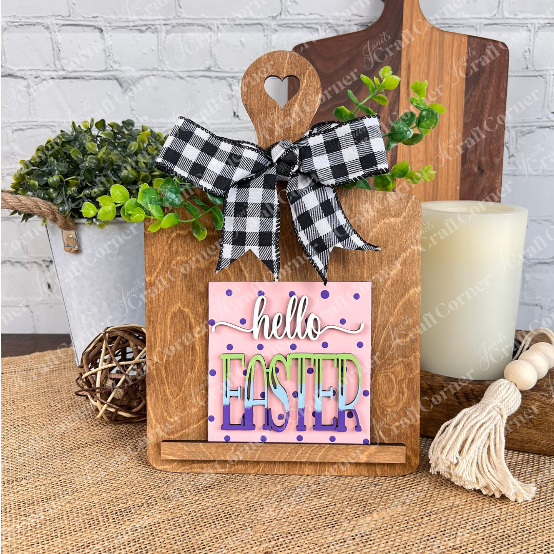 The Hello Easter Squares by Janet's Craft Corner features a wooden cutting board with a heart cutout and checkered bow, displaying "hello EASTER" in colorful text. Greenery, a vase, and coffee-themed decor complete this charming rustic look.