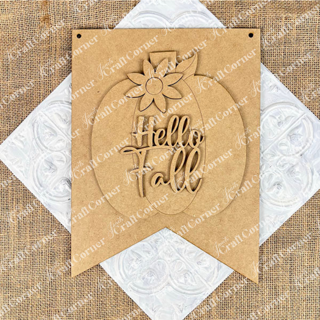 The "Hello Fall Wall Hanger" from Janet's Craft Corner features a carved pumpkin and flower atop "Hello Fall," ideal for seasonal décor on a textured white surface with a burlap background.