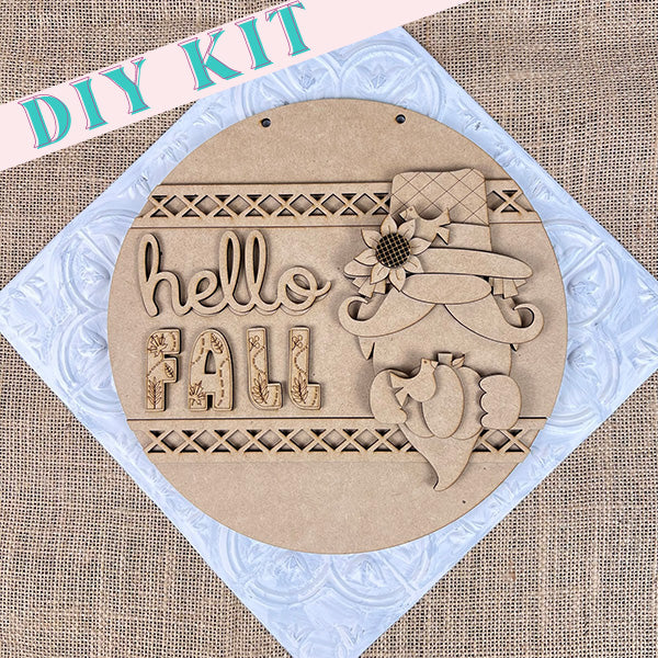 The Gnome Hello Fall Door Hanger from Janet's Craft Corner is a delightful home décor piece that features a round wooden design with the phrase "hello FALL" beside a carved gnome holding a pumpkin on textured fabric. A pink banner above identifies it as a "DIY KIT," making it perfect for adding your personal creative touch.