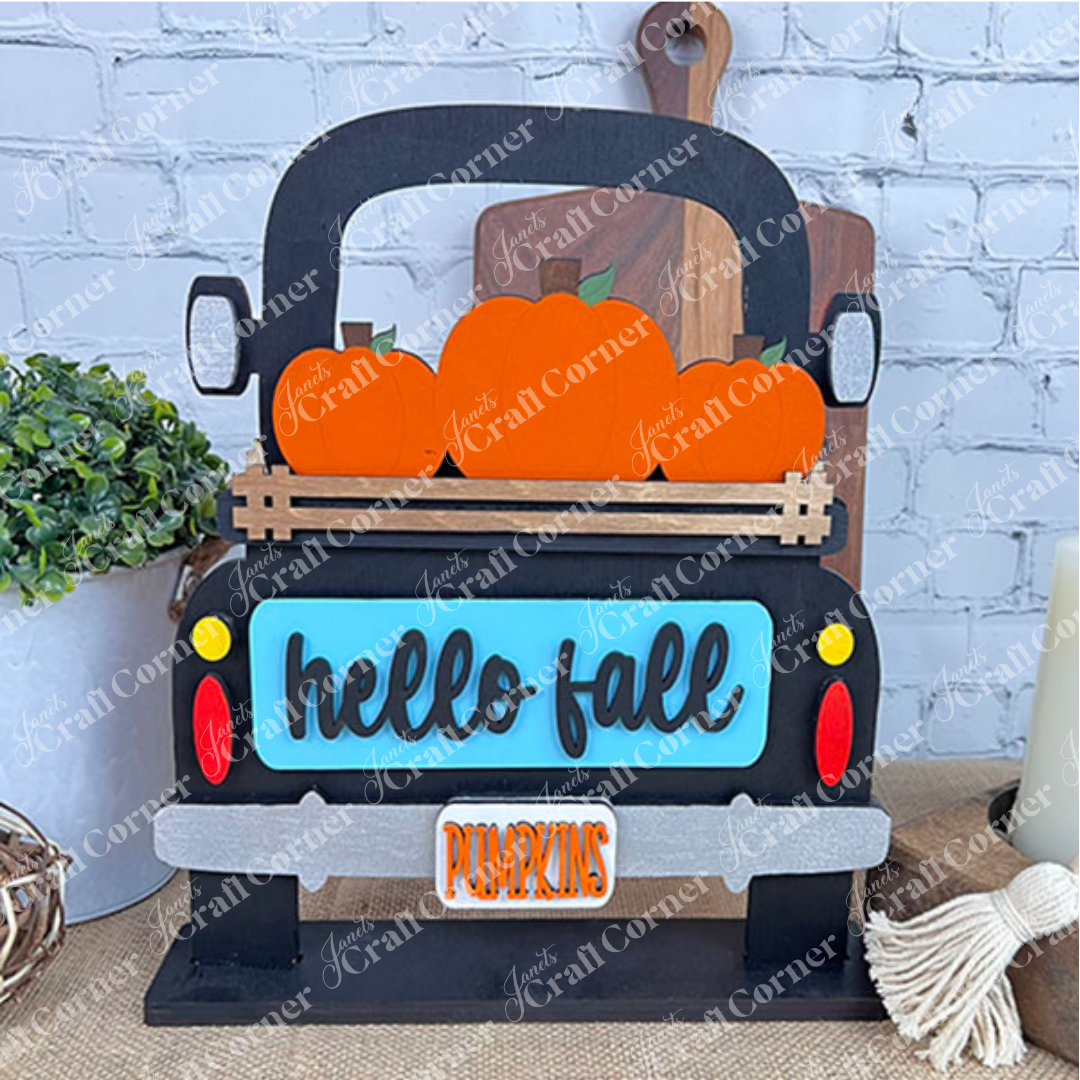 Introducing the "Hello Fall Interchangeable Insert" from Janet's Craft Corner—a charming DIY home decor kit featuring a sign shaped like a truck bed carrying orange pumpkins. With "hello fall" inscribed on the truck's blue tailgate and a "PUMPKINS" license plate, it's perfect for seasonal decoration. Pair it with items like a potted plant, cutting board, and candle for an extra touch of charm.