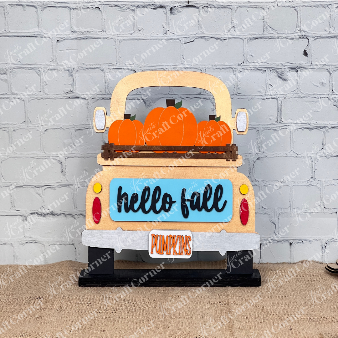 Introducing the "Hello Fall Interchangeable Insert" from Janet's Craft Corner—a charming DIY home decor kit featuring a sign shaped like a truck bed carrying orange pumpkins. With "hello fall" inscribed on the truck's blue tailgate and a "PUMPKINS" license plate, it's perfect for seasonal decoration. Pair it with items like a potted plant, cutting board, and candle for an extra touch of charm.