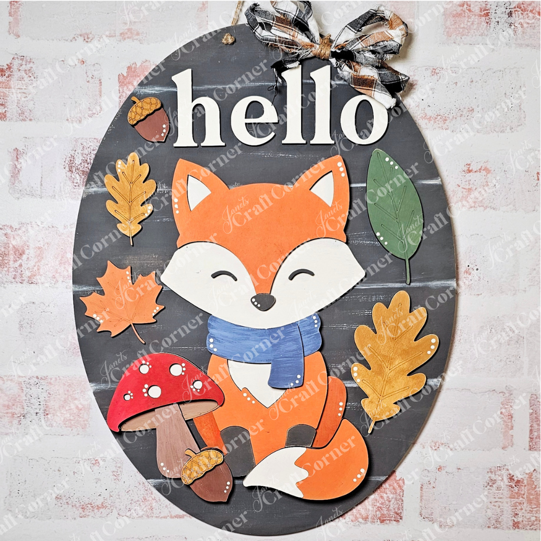 The Hello Fox Oval Door Hanger by Janet's Craft Corner is a DIY craft kit featuring a smiling fox in a blue scarf with autumn leaves and a red mushroom, topped with a plaid bow—perfect for charming seasonal home decor.