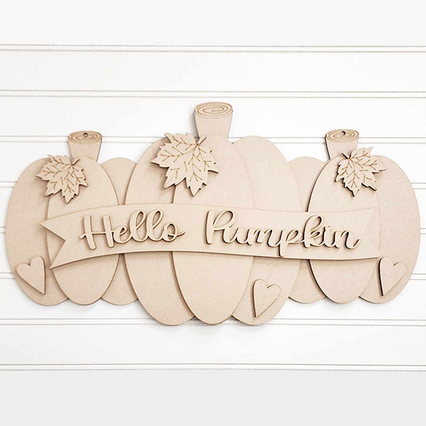 The "Hello Pumpkin Door Hanger" from Janet's Craft Corner, part of a DIY home decor kit, includes a decorative wooden sign featuring three pumpkins with leaves and the message "Hello Pumpkin" on a banner. It is adorned with two small hearts at the bottom and set against a white panel wall, making it perfect for welcoming guests to your home.